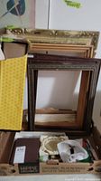 Assorted picture frames stacked against a wall with a yellow polka-dot sleeve and various other household items nearby.