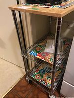 Side angle showing wood top, chrome frame, three shelves with floral liners, and rolling castors.