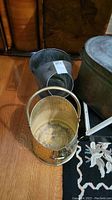 Hammered brass fireside wood bin and galvanized steel coal scuttle shown on floor next to a rug and furniture.