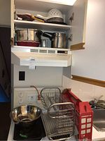 Overall view showing pots, pans stacked in upper cabinet, metal dish drying rack on counter with drip tray and utensil holder, stainless steel mixing bowl, and some cooking utensils.