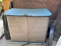 Front view of Rubbermaid plastic garden storage cabinet with double doors and curved green top lid. Shows weathering and some damage on one side door hinge.