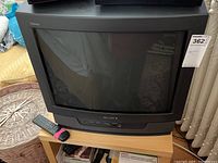Front view of Sony Trinitron CRT TV with remote control on wooden stand, showing screen and brand label.