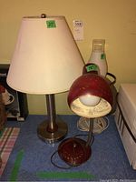 Three table lamps side by side including tall metal base lamp with beige shade, vintage red desk lamp with perforated design, and glass base lamp with brass accents.