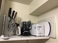 Photo of shelf contents showing acrylic knife block with 3 black handled knives and 1 wooden handled knife plus white handled scissors, mini food processor and white toaster with dark blue trim next to white cutting boards.