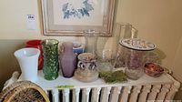Photo showing a shelf with a variety of vases including green textured, white opaque, purple, beige ceramic with handles, clear glass vases and smaller decorative pieces.