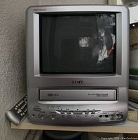 Front view of the RCA 26/32 micron VHS TV video system showing screen, VHS tape slot, buttons, and AV inputs with remote holder on left side.