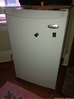 Full front view of white Whirlpool bar fridge sitting on the floor under a window with a power cord connected to the back.