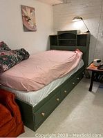 Green stained wooden captain's bed with 3 storage drawers under the bed and bookshelf headboard with shelves, shown with pink fitted sheet and lamp on headboard shelf.