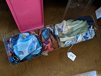 Two clear plastic shoebox-sized bins filled with assorted silk scarves in various colors and patterns on carpet floor.