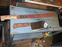 Photo showing both vintage tools - machete with original red paint and wooden handle, and heavy kitchen cleaver with wooden handle.