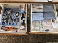 Two kitchen drawers with neatly arranged stainless steel flatware including knives, forks, spoons, and serving utensils, along with various small household items and kitchen linens.
