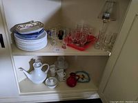 Two-shelf cupboard with porcelain coffee set on lower shelf and glassware and milk glass plates on upper shelf.