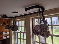 Kitchen utensils hanging from a ceiling pot rack system, including strainers, colanders, and sieves, displayed in a kitchen window setting.
