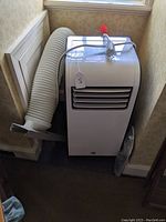 White Kenmore portable air conditioner positioned near wall with attached white flexible exhaust hose and metal window vent kit.