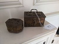Photo showing two pierced brass boxes with visible latch locks and lid handles, placed on a white surface against white paneling.