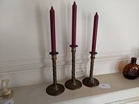 Front view of three brass candlesticks arranged in graduated height holding burgundy candles