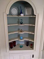 Full view of built-in shelf with various decorative glass pieces arranged on four shelves, including milk glass, colored glass tumblers, and art glass sculptures.