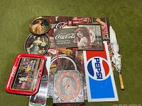 Overview of Coca Cola memorabilia including puzzle, trays, clock, posters and umbrella on green surface.