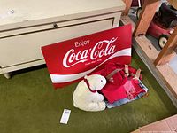 Overview of Coca Cola sign, clothing items, and white plush teddy bear on green carpet.