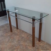 Side angle showing the glass top on wooden cylindrical legs with chrome caps and a horizontal chrome bar between the legs.