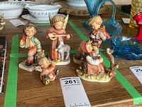 Five Hummel figurines arranged on a wooden surface, showing detailed painting and naturalistic poses of children and animals including an angel playing an instrument, a boy with a backpack, and a girl sitting with a bird.