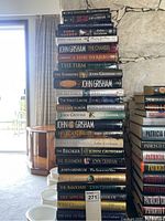 Photo showing a tall stack of John Grisham novels including a mix of hardcover and paperback books, titles visible include The Testament, The Innocent Man, The Runaway Jury, The Associate, The Appeal, The Confession, and others.