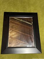 Single rectangular mirror with broad black frame lying flat on green carpet.