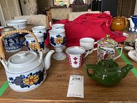 View of tea pots and cups on wooden table, showing floral and geometric patterned cups and multiple teapots including woven handle and green teapot
