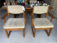 Two oak upholstered chairs side by side showing beige fabric seats and backs with zigzag pattern; visible wear and fabric damage on upholstery; wooden armrests and legs visible