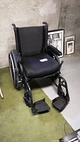 Black Quickie wheelchair positioned on a green carpet against a concrete wall, showing the front and left side with footrests extended.