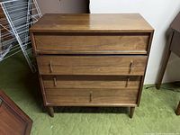Front view of mid-century three-drawer dresser showing wood grain finish, integrated handles and tapered legs