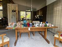 Solid oak wood table shown with various items on top in a living room setting, carpeted floor, chairs surrounding, and windows with curtains in background.