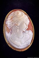 Front view of oval 14kt yellow gold cameo brooch pendant with detailed carved female profile in white relief on reddish-orange background.