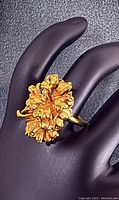 Front angled view of the gold-tone flower ring showing detailed petals on a black mannequin hand.