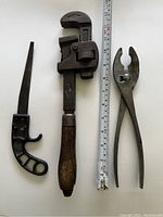 Three antique tools including a pipe wrench with wooden handle, air blow gun, and pliers alongside a measuring tape showing length