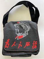 Front view of black handbag with Mao Zedong portrait and red Chinese lettering on flap, showing shoulder strap.