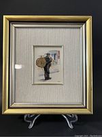 Framed painting of a one-man band musician with drum, cymbal, and pipe, shown on stand against black background.