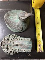 Photo of two bronze items: a bird figure and a carved ornament with intricate detailing, measuring tape visible for size reference