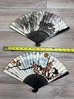 Opened view of two Chinese folding fans showing painted designs, measured against tape for scale.