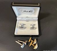 Three pairs of cuff links displayed, including one pair in original Colibri of London box and two pairs (one silver-tone, one gold-tone) outside the box on a black surface.