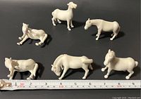 Six white porcelain animal figurines shown on black surface with measuring tape. Animals include horses and cattle in different poses, sizes around 2 to 3 inches tall and 2 inches wide.