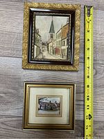 Two framed paintings side-by-side on wood floor with measuring tape showing sizes.
