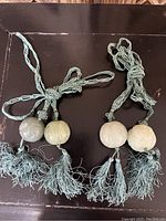 Two antique green jade spherical carved units attached to pale green braided cords with tassels shown on a dark surface.