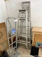 Two aluminum step ladders and hand truck against wall