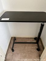 Black adjustable under-bed table positioned against wall showing overall frame and wheels.