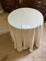 Photo of round glass topped decorator table covered with beige lightweight fabric tablecloth, showing three legged structure and measurements.