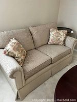 View of one beige/gold tone loveseat with damask pattern fabric and two floral patterned throw pillows stacked on it.