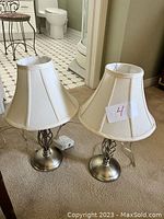 Two antique finish silver metal table lamps with white fabric shades on carpet