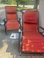 Two orangey red zero gravity chairs side by side, showing adjustable headrest pillows, sling fabric seats, powder coated steel frames, and one built-in side table with cup holder