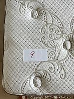 Close-up photo showing the quilted fabric corner of the mattress with decorative swirl pattern and tufted buttons; includes a lot number sticker '9'